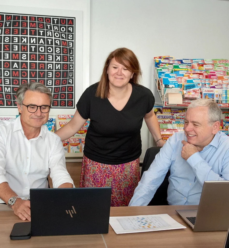 Three colleagues smiling and working together on laptops in a colorful office with magazines and a word puzzle poster on the wall.