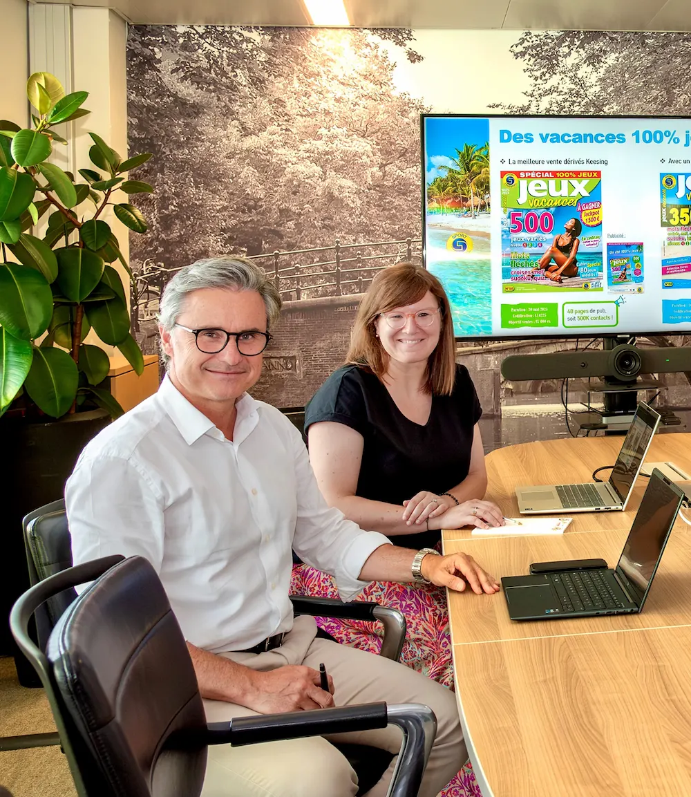 Two colleagues sitting at a meeting table with laptops in front of a presentation screen displaying a French holiday-themed slide.