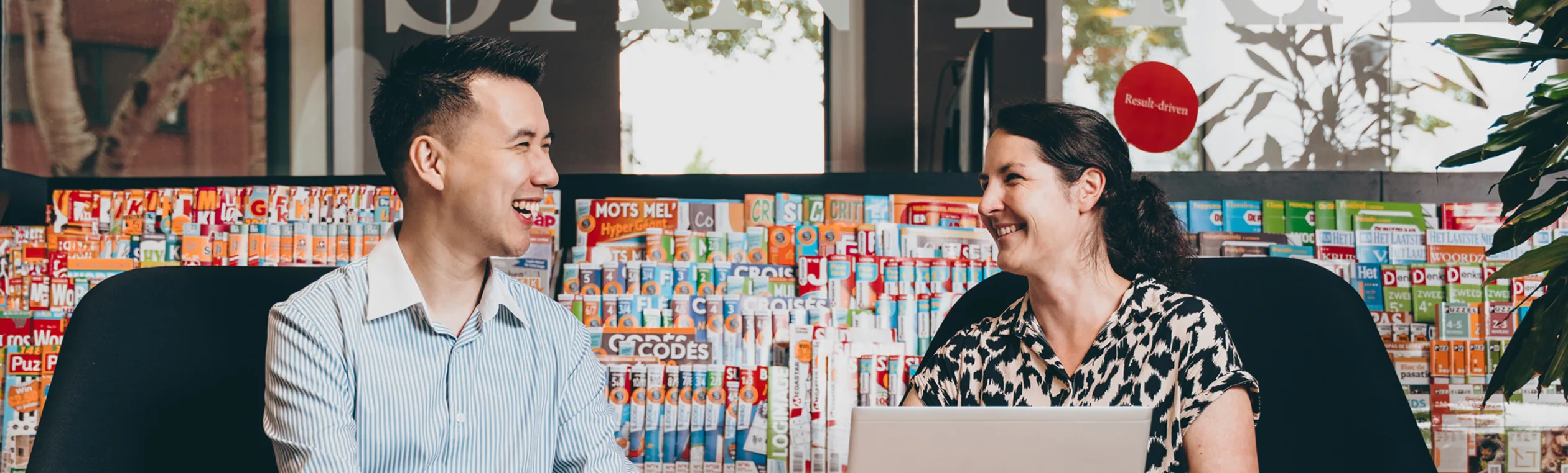 Two colleagues smiling and talking in front of a wall filled with colorful magazines.