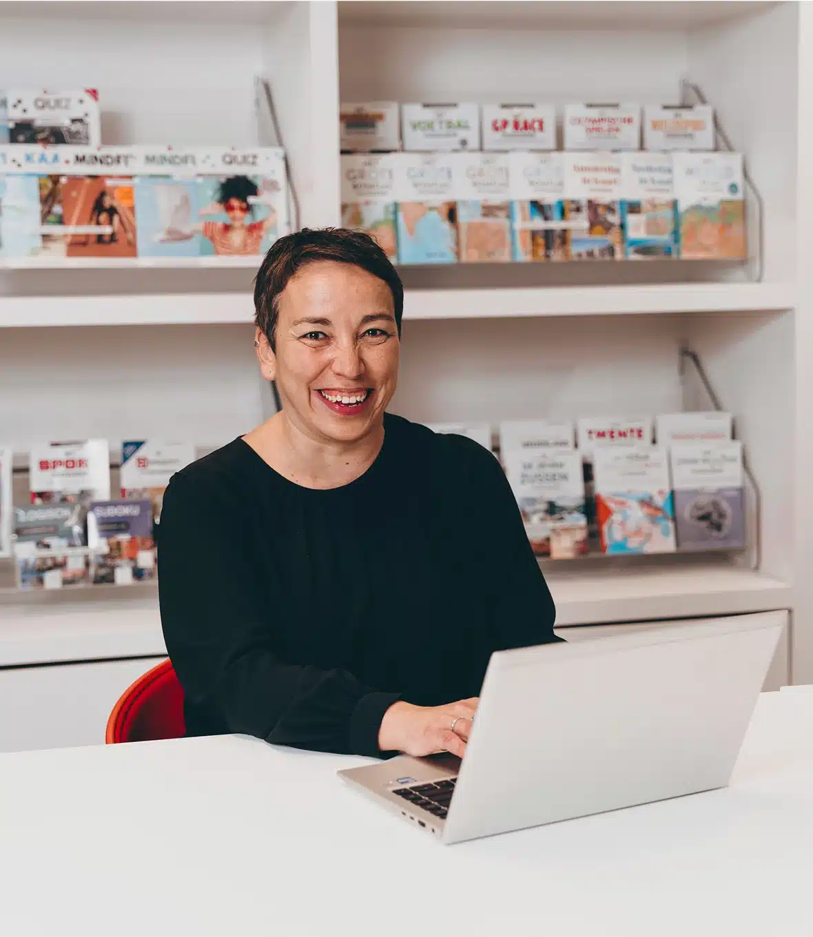 Smiling woman sitting behind a laptop in front of a wall with travel brochures.