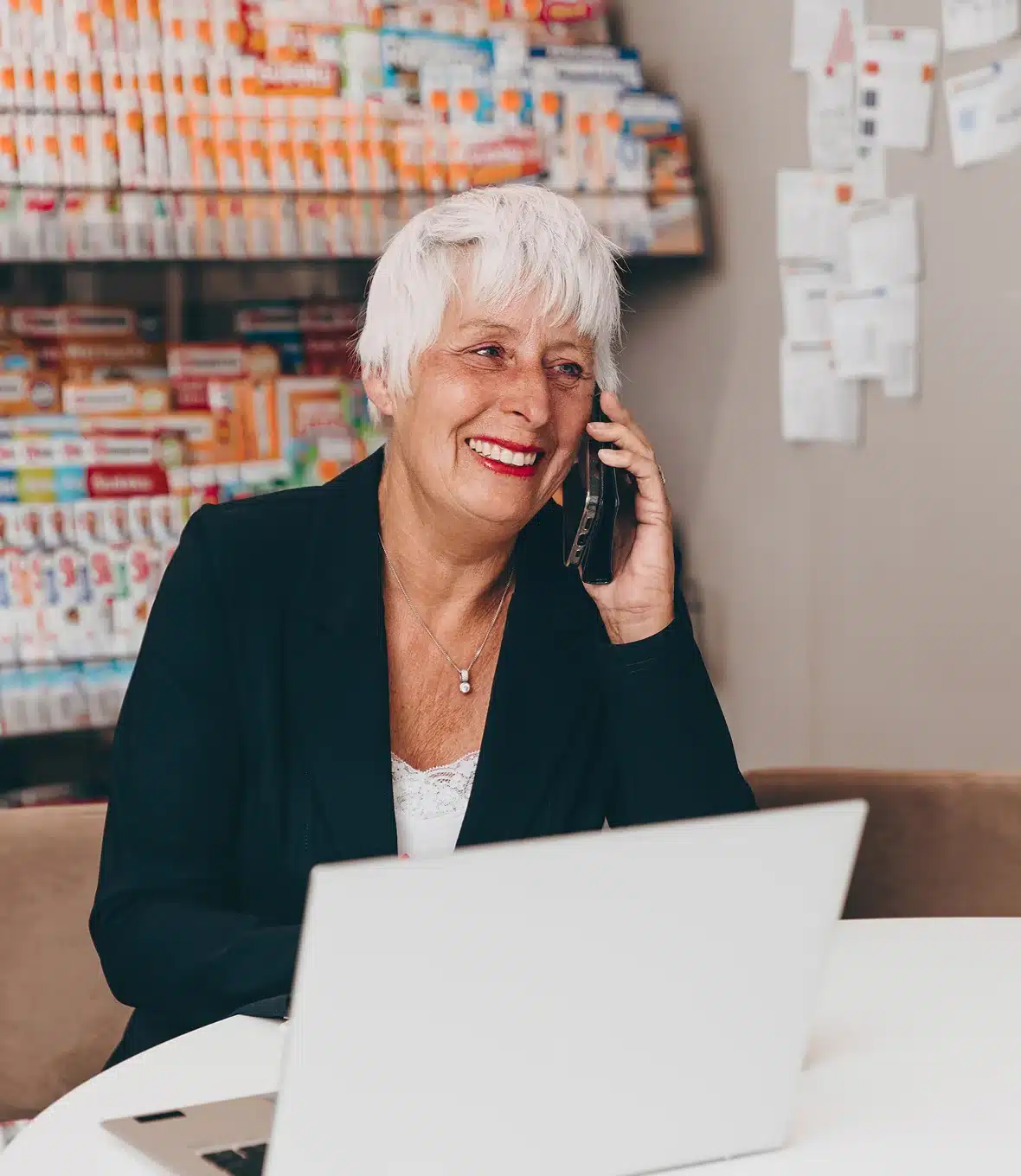 Smiling woman with short white hair talking on the phone while sitting at a desk with a laptop in front of pharmacy shelves.