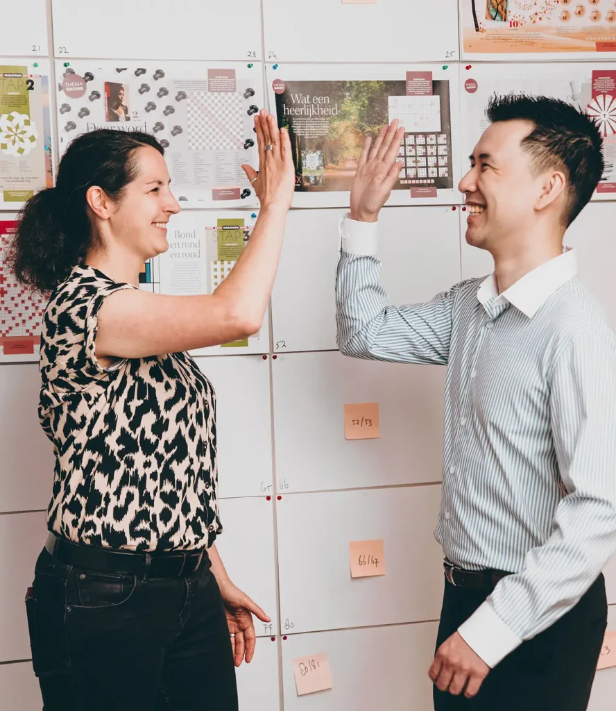 Two colleagues high-five in front of an idea board filled with printed layouts and sticky notes.
