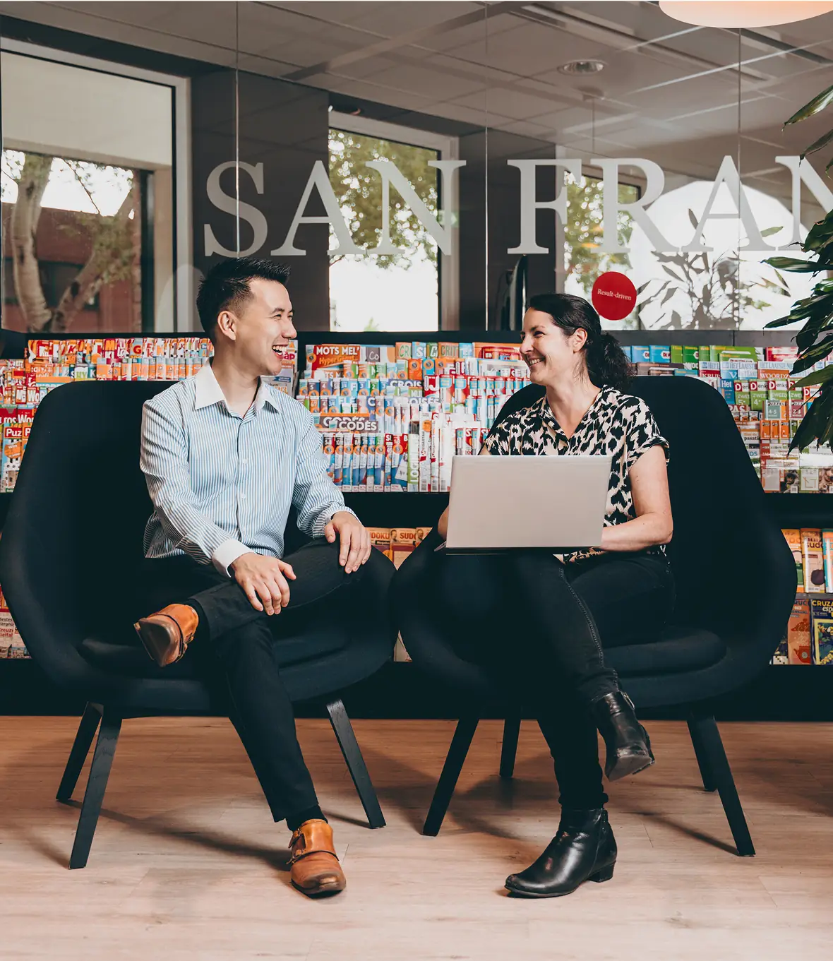 Two colleagues sitting in black chairs having a friendly conversation, with a laptop and colorful magazine display in the background.