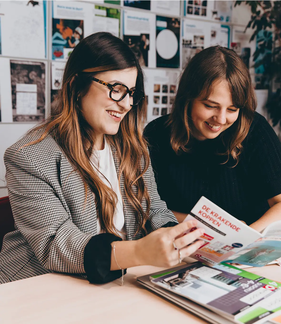 Two women smiling while reading a magazine together at a desk.