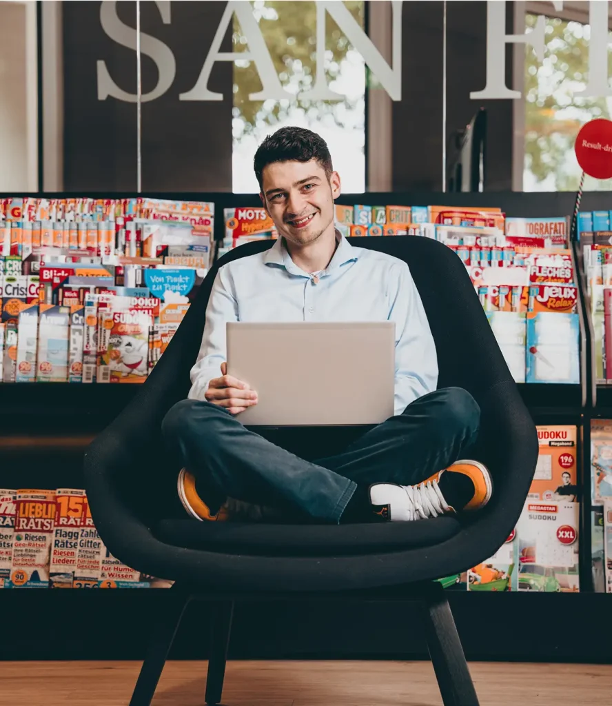 Man sitting cross-legged in a black chair with a laptop in front of a magazine display.